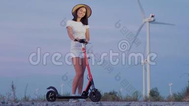 黄昏时分，在风力发电机的背景下，女孩戴着一顶草帽坐在滑板车上。