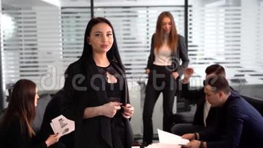 办公室里微笑的女商人，双手交叉，自信的表情，其他员工在办公室开会