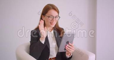 一位戴眼镜的年轻白种人女商人的特写肖像，他在平板电脑上打着一个视频电话，坐在平板电脑里打招呼