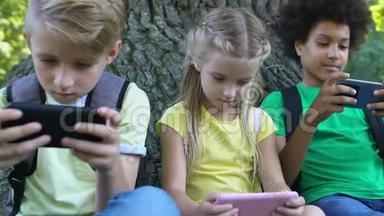 孩子们坐在公园的树下玩智能手机游戏，玩电子游戏成瘾