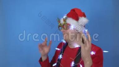 穿着圣诞老人服装和新年灯的年轻白种人正在蓝色背景上跳舞。 新年假期