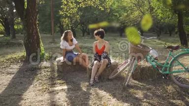 两个积极的女人在夏天的花园里坐在干草堆上说话，她们的自行车站在附近。 农村生活