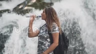 迷人的女孩从瓶子里喝冷水，当站在瀑布附近的石头上时，解渴。 侧面