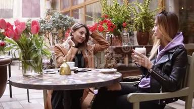 两位迷人的女士在舒适的户外咖啡厅一起享受咖啡。 他们坐在咖啡店的桌子旁聊天