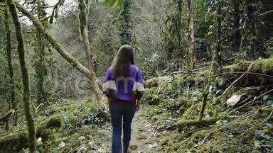 穿着<strong>运动夹克</strong>的长发女孩走过一条森林小径，穿过覆盖着绿色苔藓的神秘树木。