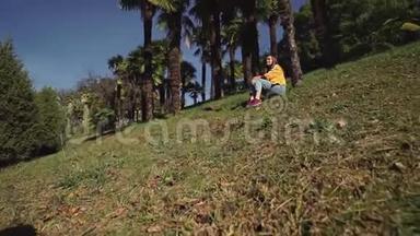 穿着牛仔裤和夹克的可爱女孩坐在棕榈树<strong>下</strong>的绿色草坪上
