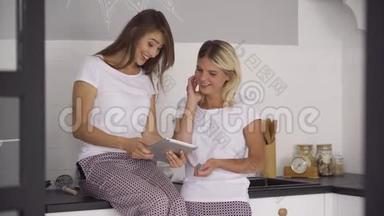 两个穿着同一件睡衣的快乐女朋友一起在家里的厨房里用平板电脑。 沉迷于