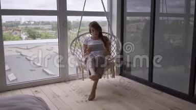 苗条的年轻女子在一张挂着的椅子上摆动，同时在大的<strong>落地</strong>窗上使用她的平板电脑。 自由职业者