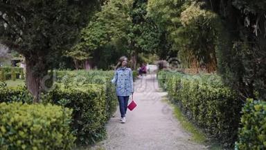 女孩沿着剪好的绿色灌木丛走在公园里，把她的手放在树叶上，总计划