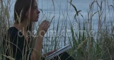 美丽的白种人黑发女孩坐在湖岸上，带着书，看着远方。 一个美丽的女人
