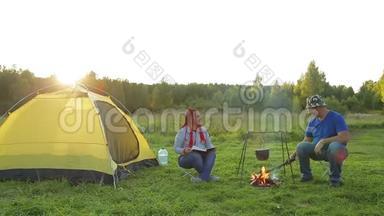 一个男人和一个女人在篝火附近的帐篷附近读一本书，看<strong>沸腾</strong>的<strong>锅</strong>。