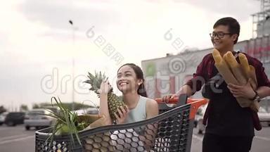 可爱的年轻亚洲女人坐在购物车里，手里拿着纸袋，把她英俊的男朋友推着吃东西