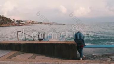 孤独悲伤的女孩看着大海。 站在码头的孤独的年轻女人。 孤独的女孩在恶劣的天气散步。 少女