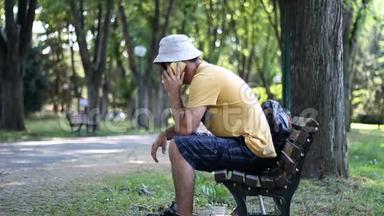 戴着帽子和太阳镜的人坐在公园长凳上打电话。 在树下的公园里放松时，男人用电话交谈