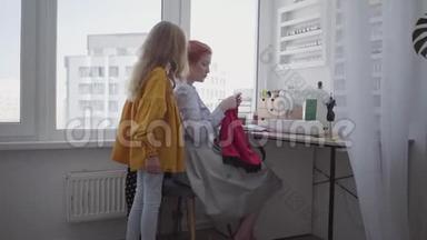 美丽的红发女人缝衣服坐在桌子旁边的窗户在灯光室。 小女儿看上去