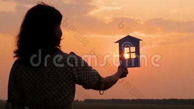 一个穿着裙子的女孩拿着纸房子的剪影。 夜晚美丽的日落。 家庭幸福和舒适..