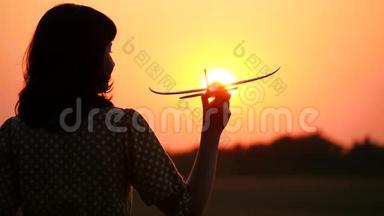 一个女孩的剪影，她拿着一架模型飞机面对美丽的日落。 旅游和航空旅行。 梦想和梦想