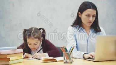 年轻的母亲坐<strong>在</strong>桌旁时<strong>在</strong>笔记本电脑上工作，女儿<strong>在此</strong>期间写作业。 灰色背景。