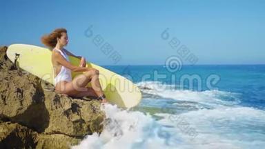 冲浪女孩坐在一个美丽的岩石海滩与木板。 强<strong>大</strong>的<strong>海浪</strong>冲击着岩石海岸。 女孩看着远处。