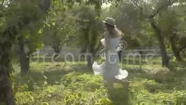 可爱的年轻女子戴着草帽，穿着白色的长裙，穿过绿色的夏日花园。 免费农村生活