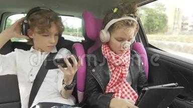 男孩和女孩带着耳机在汽<strong>车上</strong>玩平板电脑和智能手机，孩子们在汽<strong>车上</strong>使用设备。 兄弟和兄弟
