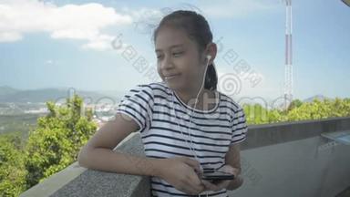 年轻的亚洲女孩愉快地从手机里听音乐，站在风景优美的观景塔上。