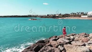 穿红色衣服的女人走在岩石海滩上，举起双手分开在空中看着蓝色的海洋和天空。 风在拍打她的裙子。