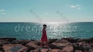 穿着红色裙子的可爱女士在海上码头的岩石上随风拍打，蓝色海水上有阳光反射。 天气晴朗炎热