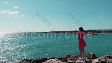 穿着红色裙子的女人，双手分开，迎着阳光站在岩石悬崖码头，享受着风和阳光。 海浪撞击岩石