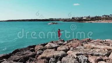 双手分开的女人穿着红色可爱的裙子，风<strong>拍打</strong>着站在岩石悬崖上，看着太阳路和蓝色的<strong>海水</strong>。