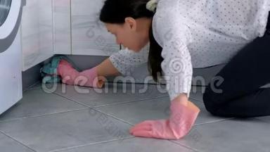 穿着粉红色橡胶手套的女人用布洗硬厨房家具。 坐在地板上。 侧视。