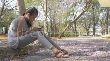 穿着休闲装的亚洲可爱女孩坐在树下，喜欢在公共公园里用手机带耳机。