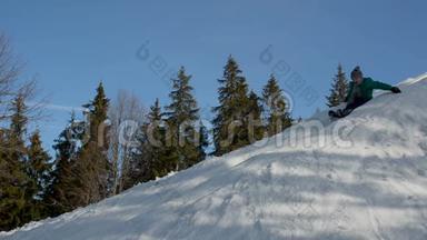 穿绿色夹克的男孩从高高的雪山上滑下来。 冬日阳光明媚。 蓝天和云杉背景