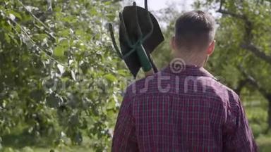 年轻农民肩上扛着一把铲子和一把草叉走过花园的后景。 <strong>农村生活</strong>观念