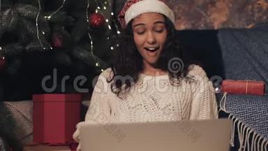 美丽的年轻女孩戴着桑塔帽坐在<strong>圣诞树</strong>下使用她的笔记本电脑。 打<strong>视频</strong>电话的女孩