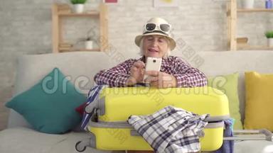 一个戴帽子的老女人微笑着，手里拿着旅行箱的智能手机坐在沙发上