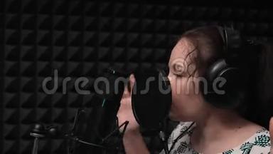 漂亮的女孩戴着蓝眼睛的耳机在录音室的麦克风附近唱歌。 录音。 激情歌唱
