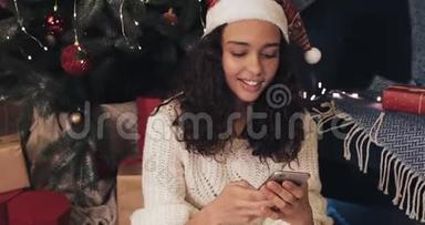 一个笑容可掬的年轻女孩在圣诞树附近的现代手机上发短信，在社交网络上聊天