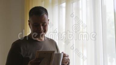 独自一人穿着<strong>卡其色</strong>的t恤站在窗边，拿着一张木框里的照片。 他检查照片和