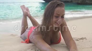 迷人的女人穿着比基尼躺在沙滩上，微笑着拍摄蓝色的海洋景观。 美女模特
