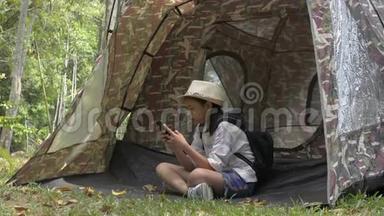 亚洲可爱的女孩，背包坐着，在自然公园的帐篷前使用手机。
