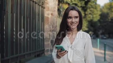 年轻迷人的微笑着的黑发女孩，在舒适的街道上，一边使用手机，一边在社交网络上聊天