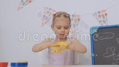 一个小可爱的女孩坐在孩子们的桌子旁`用手指画画，把她的手染成黄色