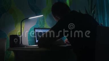 他坐在桌子前写字，在笔记本电脑前。 晚上，晚上，公寓台灯