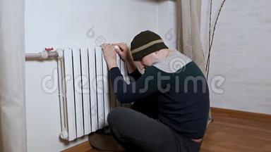 一个戴帽子的人把手放在靠近墙壁的暖气片上取暖。 公寓里冷，<strong>供暖</strong>系统差