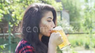 年轻快乐的女人在花园里烧烤时，用玻璃喝啤酒，解渴