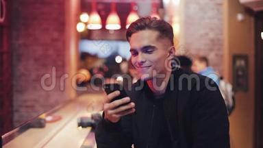 年轻迷人的男人带着美丽的微笑在晚上使用智能手机。 他坐在靠近霓虹灯的酒吧或餐馆里