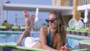 时尚迷人的女孩，戴着太阳镜和泳衣，在昂贵的度假胜地的泳<strong>池边</strong>休息在躺椅上