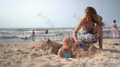 穿比基尼的母亲把她的金发小女孩埋在沙滩上。摄像机运动
