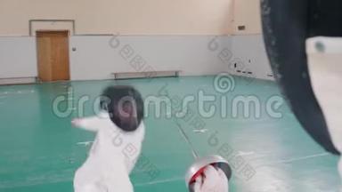 两名身穿白色防护服的年轻女子在健身房进行击剑训练-一名女子进攻，另一名女子防守
